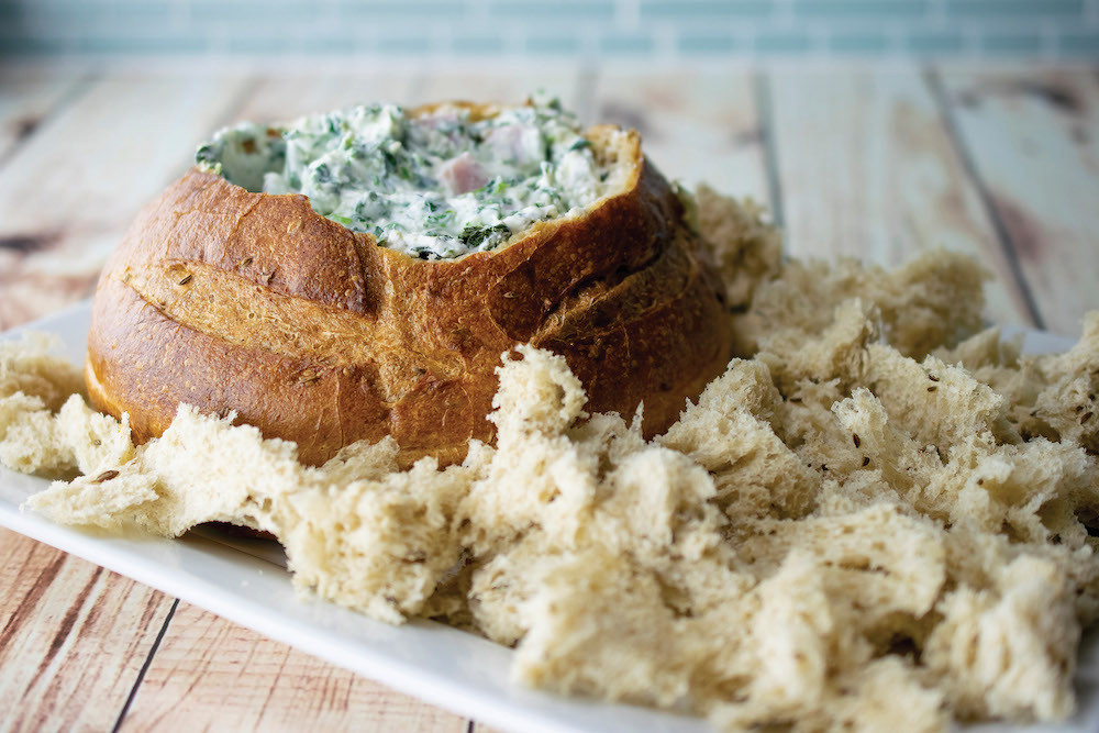 spinach and ham dip in a bread bowl with torn bread on the side