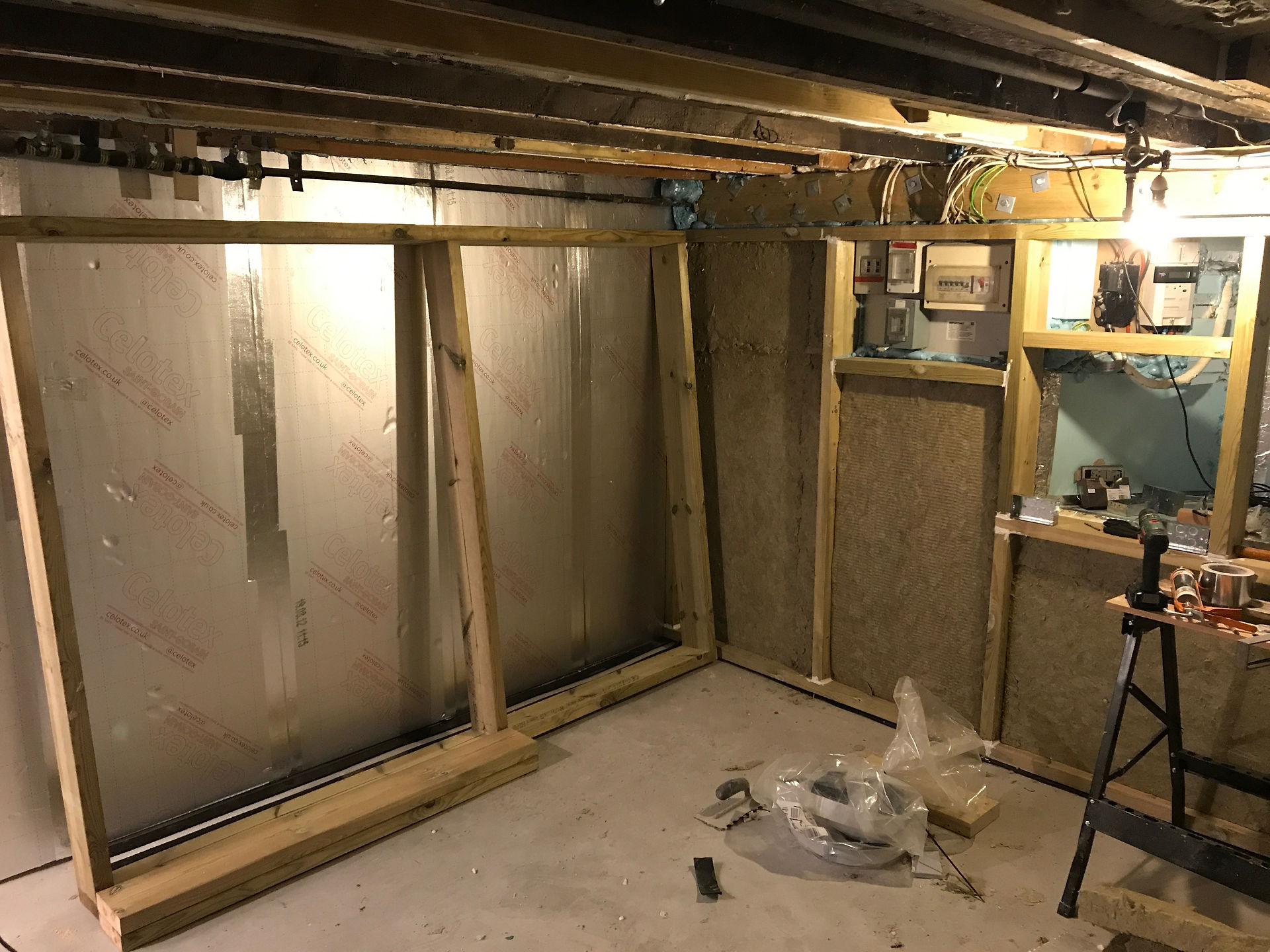 Construction site with exposed wooden beams, insulation on walls, and a lit bulb. Tools and electrical boxes on a shelf; cluttered workspace.