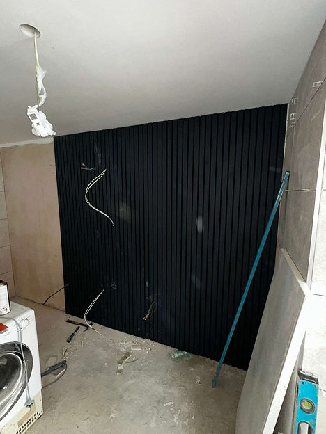 Black slatted wall with exposed wires, unfinished plasterboard, gray tiles, and a leaning blue tool in a renovation site
