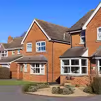A sunny suburban street featuring red brick houses with sloped roofs, a landscaped garden