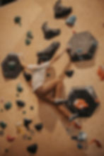 Man bouldering on an indoor climbing wall, wearing a white shirt and brown pants. Holds are multicolored; backdrop is beige plywood.