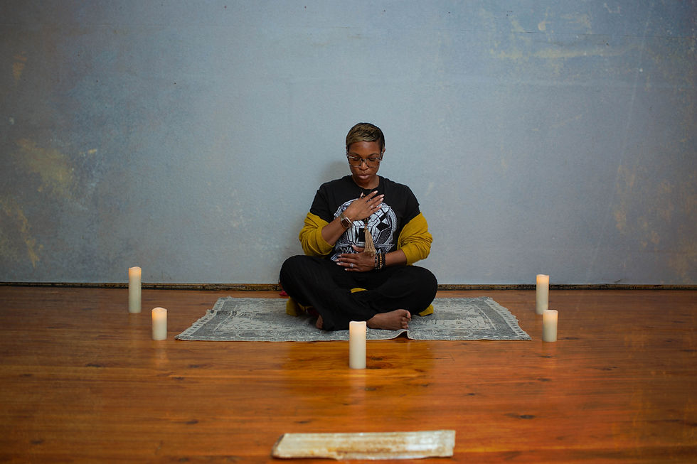 Aunty Charmaine seated cross-legged in a meditative pose on a small rug, hands resting on heart and belly, surrounded by pillar candles and a selenite wand on a warm hardwood floor.