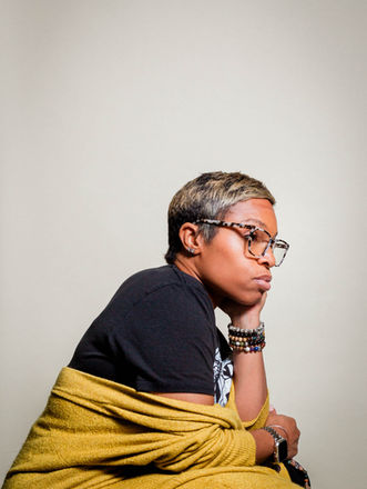 A woman sits thoughtfully with her chin resting on her hand, wearing glasses, beaded bracelets, and a mustard-yellow shawl draped over her shoulders. Her expression appears reflective and tired, as if deep in contemplation or emotional processing. The neutral background and soft lighting emphasize her quiet strength and vulnerability. In the corner, the text reads “Soul Tribe Media,” grounding the image in the context of introspection and healing — a visual companion for the OK Universe, I Hear You blog about emotional honesty and shared growth.