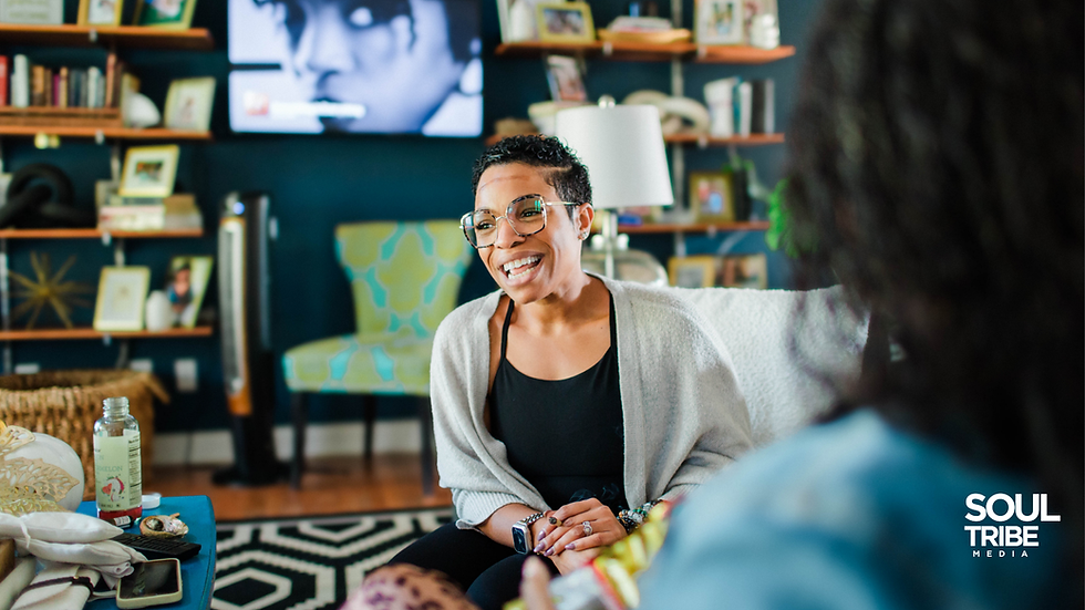 A woman smiles brightly while sitting in a colorful living room, engaged in warm conversation with another person. Books, photos, and art fill the background, giving the scene a feeling of authenticity, laughter, and shared growth — embodying the spirit of connection and belonging found in the OK Universe, I Hear You series. The Soul Tribe Media logo is shown in the corner.