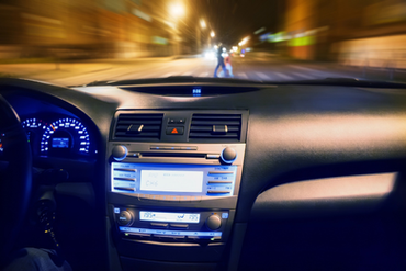 View from inside a vehicle showing the dashboard and a blurred view through the windshield of pedestrians crossing at a crosswalk at night.