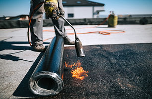 A worker wearing gloves uses a torch to melt roofing material on a flat rooftop