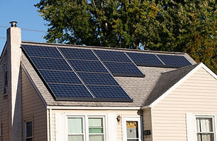 A small house with solar panels on its grey shingle roof, surrounded by lush green trees and bright sunshine