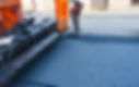 Two construction workers in bright orange uniforms and reflective vests lay fresh asphalt on a city street using a paving machine.