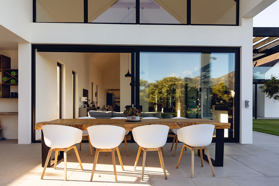 an outdoor dining area featuring a long wooden table with five white chairs arranged in front of large clean glass sliding doors. The setting highlights a modern, minimalist design with natural light, reflecting trees and a clear sky in the glass.