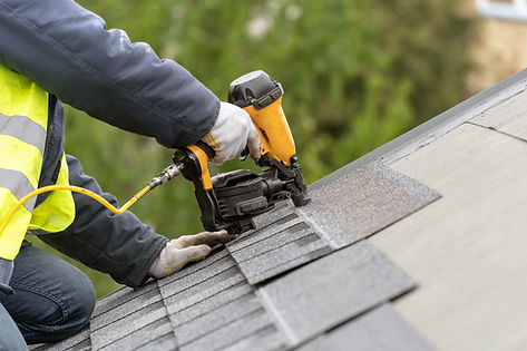 A worker in a reflective vest and gloves uses a nail gun to attach shingles to a roof