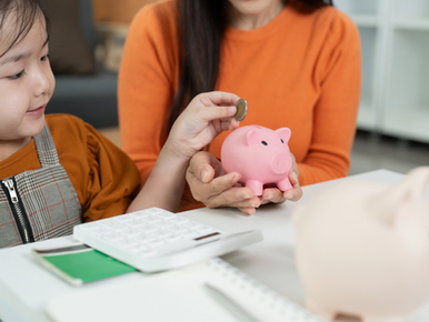 Child adding a coin to a piggy bank beside a calculator and notebook.