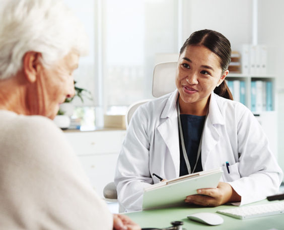 A female doctor smiles kindly at an elderly patient while holding a clipboard