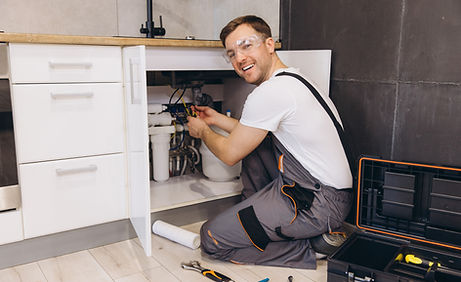 Plumber wearing safety glasses kneeling under a kitchen sink while repairing plumbing with tools beside him.
