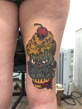 Tattoo of a cupcake with a skull and a cherry, detailed on a leg.