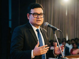 Bitan Ghosh in a suit speaks at a podium with a microphone. Audience and curtains in the background, bright spotlight overhead. Serious expression.