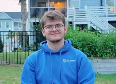Young man in blue hoodie with glasses smiles outdoors in front of a garden. A house and palm tree are in the background.
