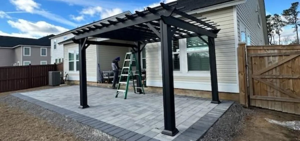 Man stands on a patio with a ladder, constructing a black pergola. House and wooden fence in the background under a blue sky with clouds.