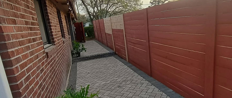 Red wood wall and wooden fence line a walkway with herringbone-patterned pavers. Green foliage at intervals; trees visible in the distance.