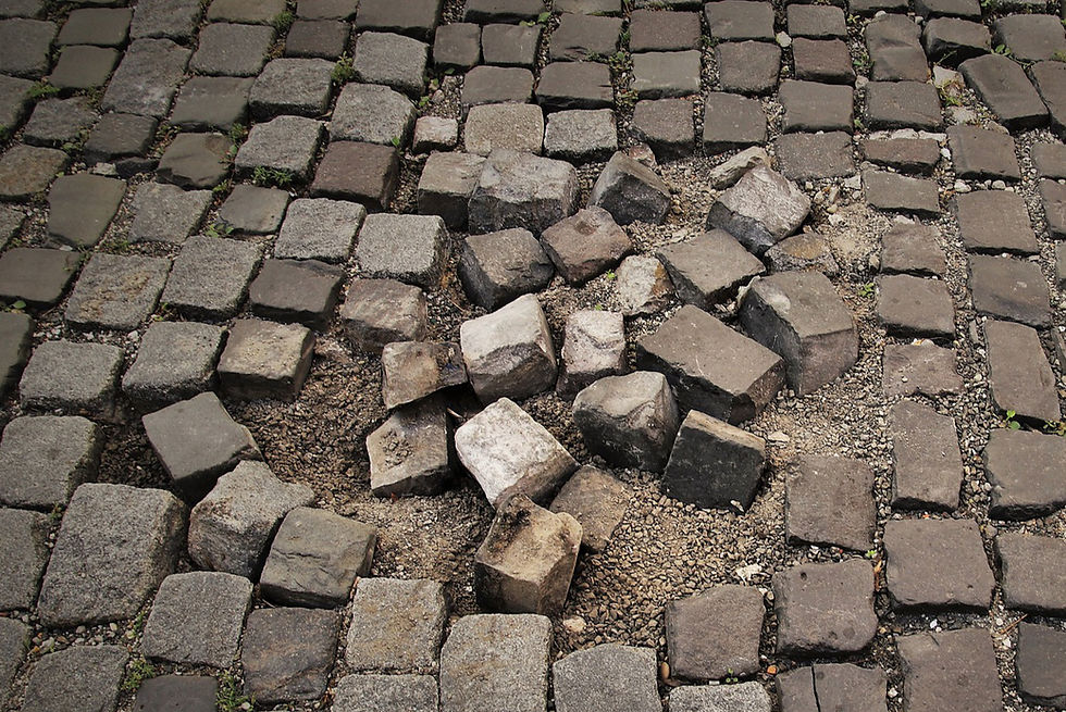 Uneven cobblestone pavement with scattered loose stones and visible gravel. Earthy tones and small green grass sprouts can be seen.