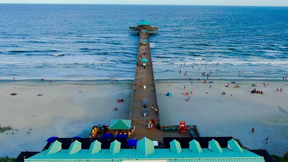 Aerial view of a beachfront with a long pier, people relaxing on white sand and swimming. Green roofs and vivid blue ocean create a tranquil scene.
