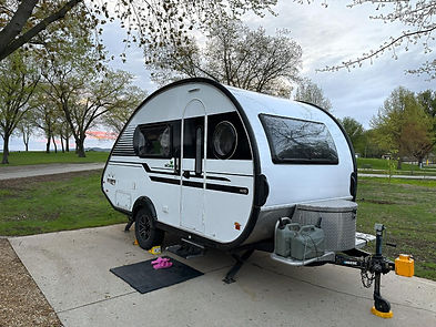 A well-maintained Corps of Engineers campground with RVs parked on clean, level sites that are slightly close together. Amenities like water and electric hookups are visible, along with nearby restrooms and shower houses. The mighty Mississippi River creates a scenic backdrop, while train tracks run along the edge of the campground, hinting at frequent train activity.