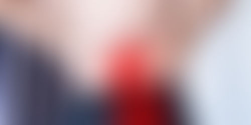 Person holds lower back with both hands, highlighting pain area in red. Wearing beige top, black pants. Blue curtain in background.