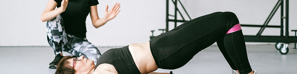 Woman in black workout clothes holds a bridge pose in a gym while a trainer kneels nearby, with equipment in background.