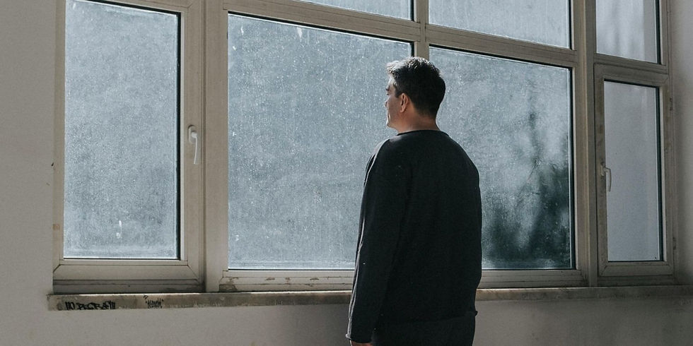 A person in a dark shirt gazes out a large, dusty window in a dimly lit room. The mood appears contemplative and somber.
