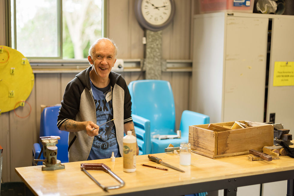 Man smiling in a workshop, holding a tool. Wooden box, saw, and glue bottle on table. Blue chairs and a clock in the background.