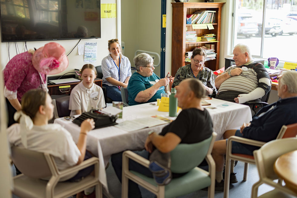 People gathered around a table in a well-lit room with a bookshelf. A woman leans in to assist.