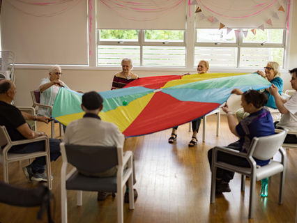 Seniors seated in a circle hold a colorful parachute in a bright room. Smiling faces, festive bunting, and wooden floor enhance the cheerful mood.