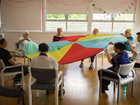 Seniors seated in a circle hold a colorful parachute in a bright room. Smiling faces, festive bunting, and wooden floor enhance the cheerful mood.