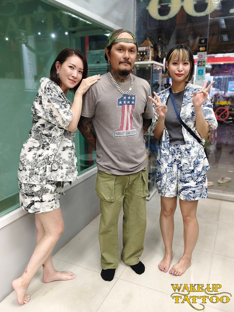 タトゥー店前で、中央の男性と左右の女性2人がポーズ。女性はピース、男性は無表情。下にWAKE UP TATTOOの文字。