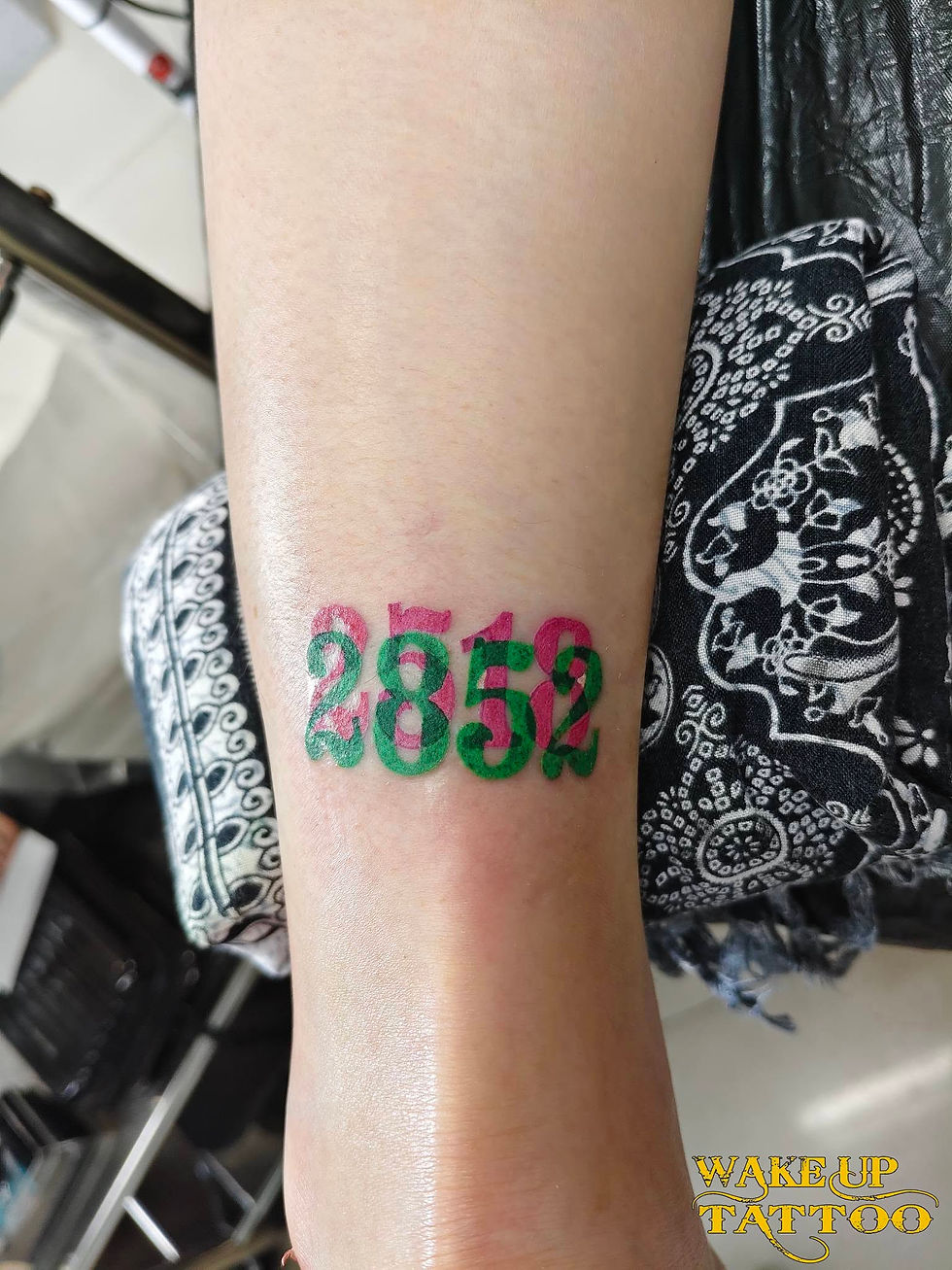 前腕にピンクと緑で重なった数字2852のタトゥー。黒白の柄布の上にあり、右下にWake Up Tattooの文字。
