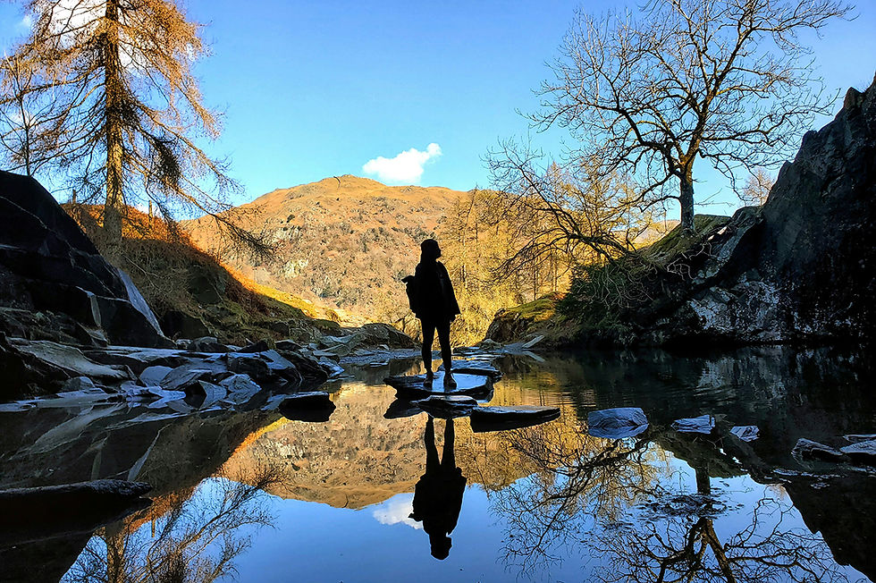 A young woman stands on stepping stones in a creek looking out over a valley in the Lake District National Park, United Kingdom. Photographer: Aniket Das on Unsplash.