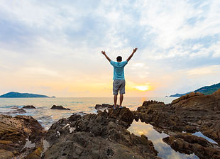 A young man celebrates his independence by standing and on a clifftop with his arms outstretched watching the sunset