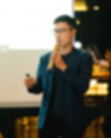 Man in a blue jacket speaks into a microphone on stage, holding a remote. Blurred background with screen showing partial text.