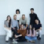 A group of eight people smiling in casual attire, sitting and standing in front of a plain white wall. One person makes a peace sign.