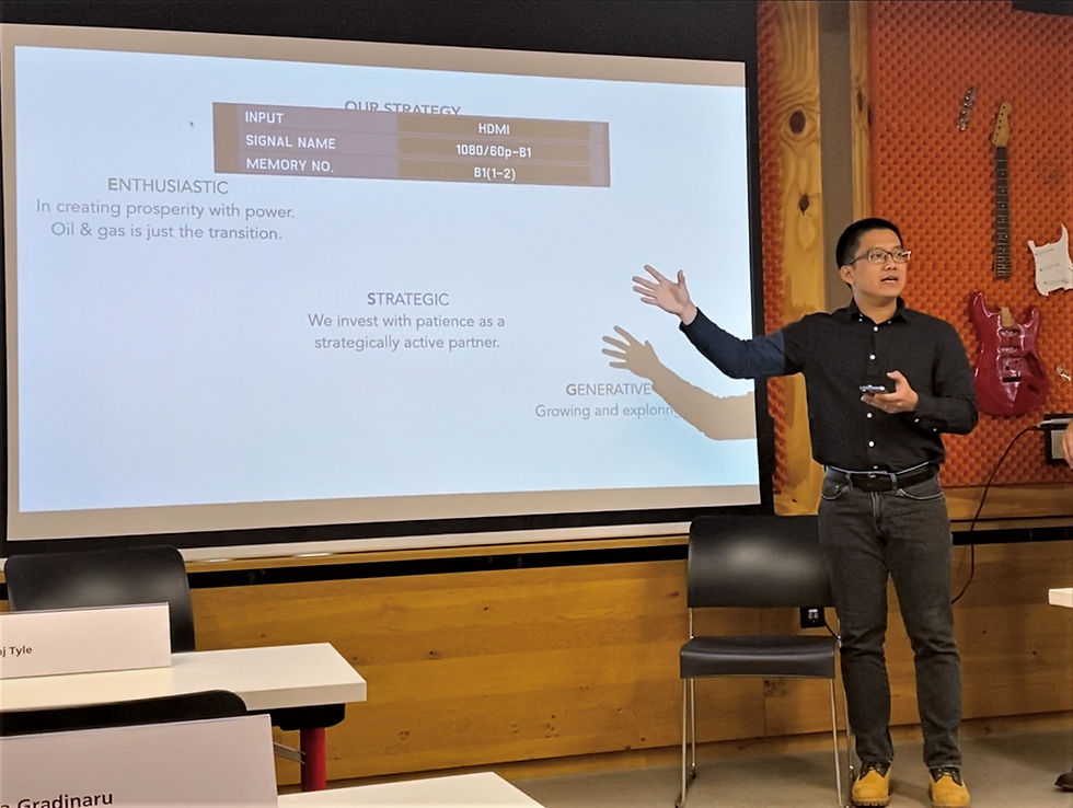 Man presenting in a room with a guitar on the wall. Projection screen displays strategic investment text. Casual setting, engaged mood.