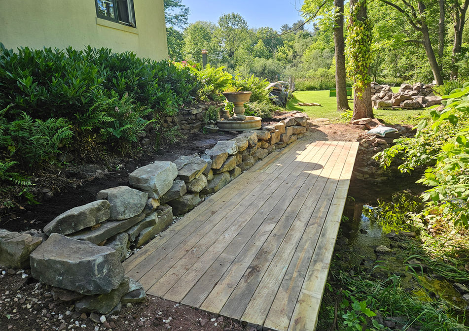 Wooden footbridge and natural stone retaining wall built by HS Premier Construction Services over a landscaped stream.