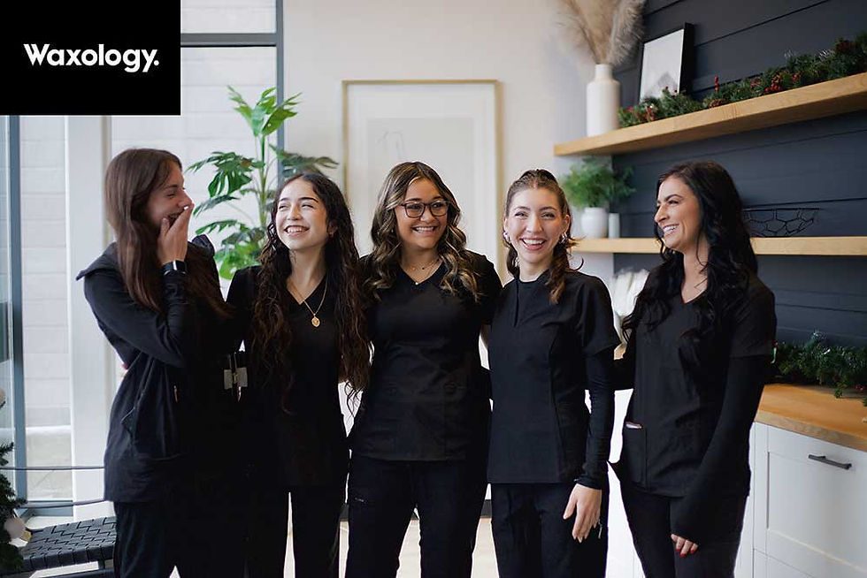 Five women in black outfits laughing and smiling in a modern room with plants and wooden shelves. "Waxology" logo visible in corner.