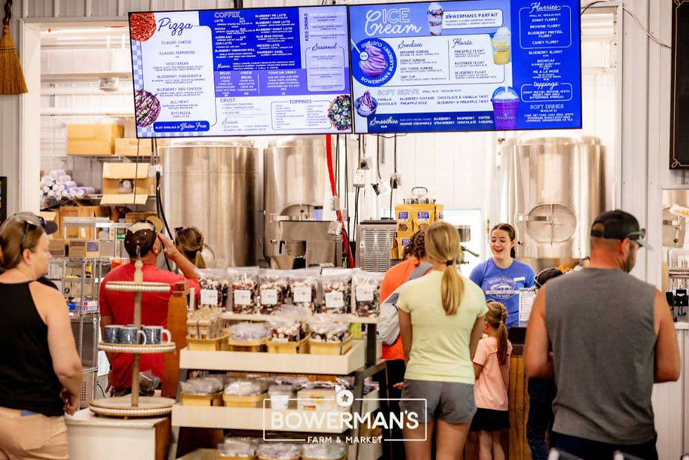 Customers order at a farm market counter with an overhead menu. A woman behind the counter smiles. Products and metal tanks are visible.