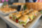 Tray of neatly arranged sandwiches with toothpicks on a counter. Background has blurred wicker basket. Text reads "Bowerman's on 8th."