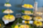 Tiered trays of lemon desserts, including cupcakes, cheesecakes, and macarons. Greenery decor, labeled "Treats" sign, blue wall.