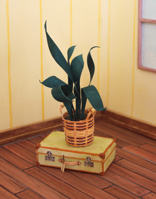 Tiny painted watercolour paper pot plant sitting in a basket on top of a suitcase on a wooden floor