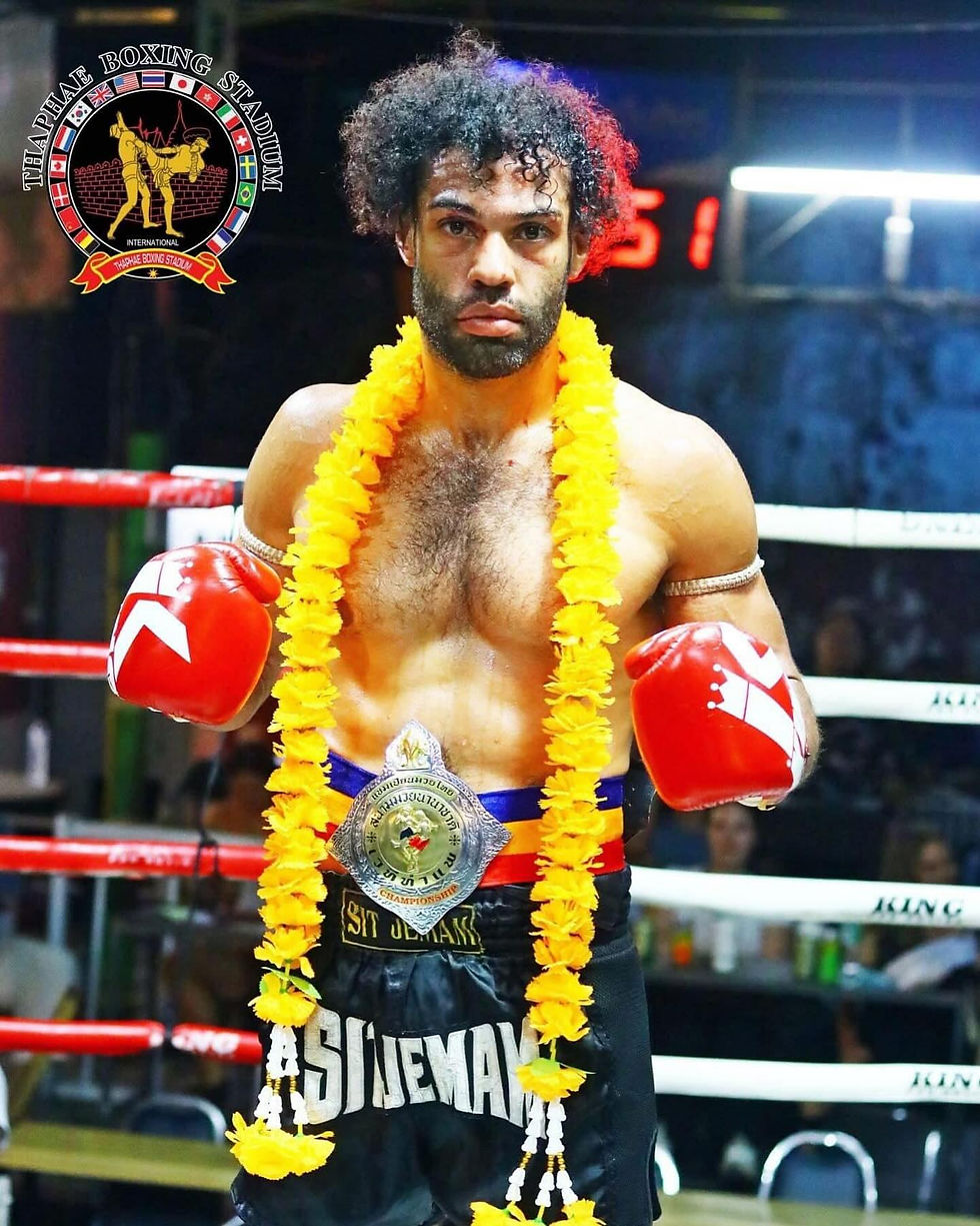Boxer with red gloves and champion belt stands in a ring. Wearing a yellow flower garland. "Thaphae Boxing Stadium" logo visible.