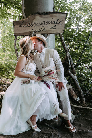 Brautpaar küsst sich in der idyllischen Naturlandschaft des KulturGut Wrechen auf einer Holzbank mit einem Schild, auf dem ‚Lieblingsplatz‘ steht – eine romantische Kulisse für ihren ganz besonderen Tag.