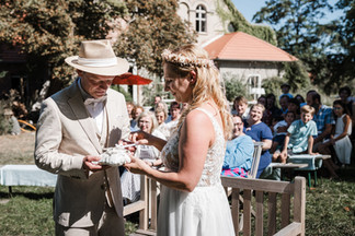 Hochzeitsfoto der freien Trauung im KulturGut Wrechen: Brautpaar öffnet das Ringkissen während der Zeremonie, im Hintergrund sind die Gäste der Trauung zu sehen.