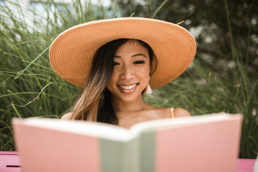 Smiling woman in a wide-brimmed straw hat reading a book outdoors. Green foliage in the background. Bright and relaxed atmosphere.