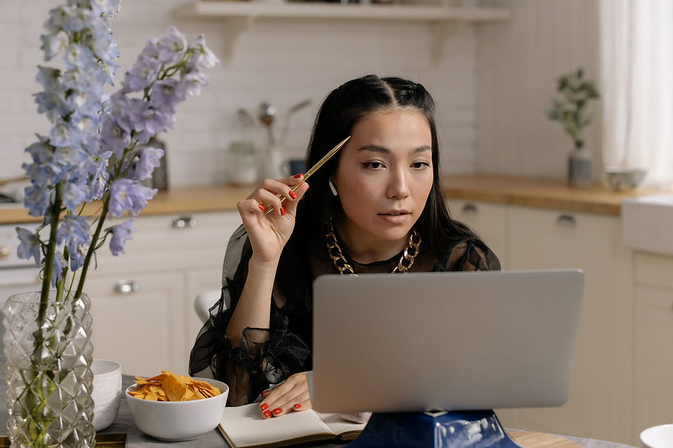 Woman with a pen looks at a laptop in a kitchen, surrounded by flowers and snacks. She appears focused, wearing a black top and gold necklace.